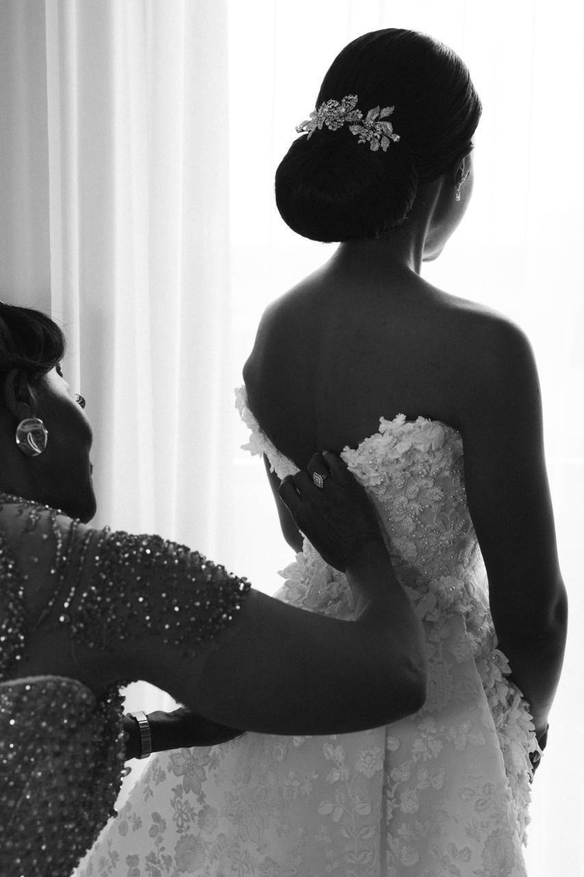 Black and white view from behind as a bride is fastened into a floral-appliquéd gown, a jewelled comb in her hair