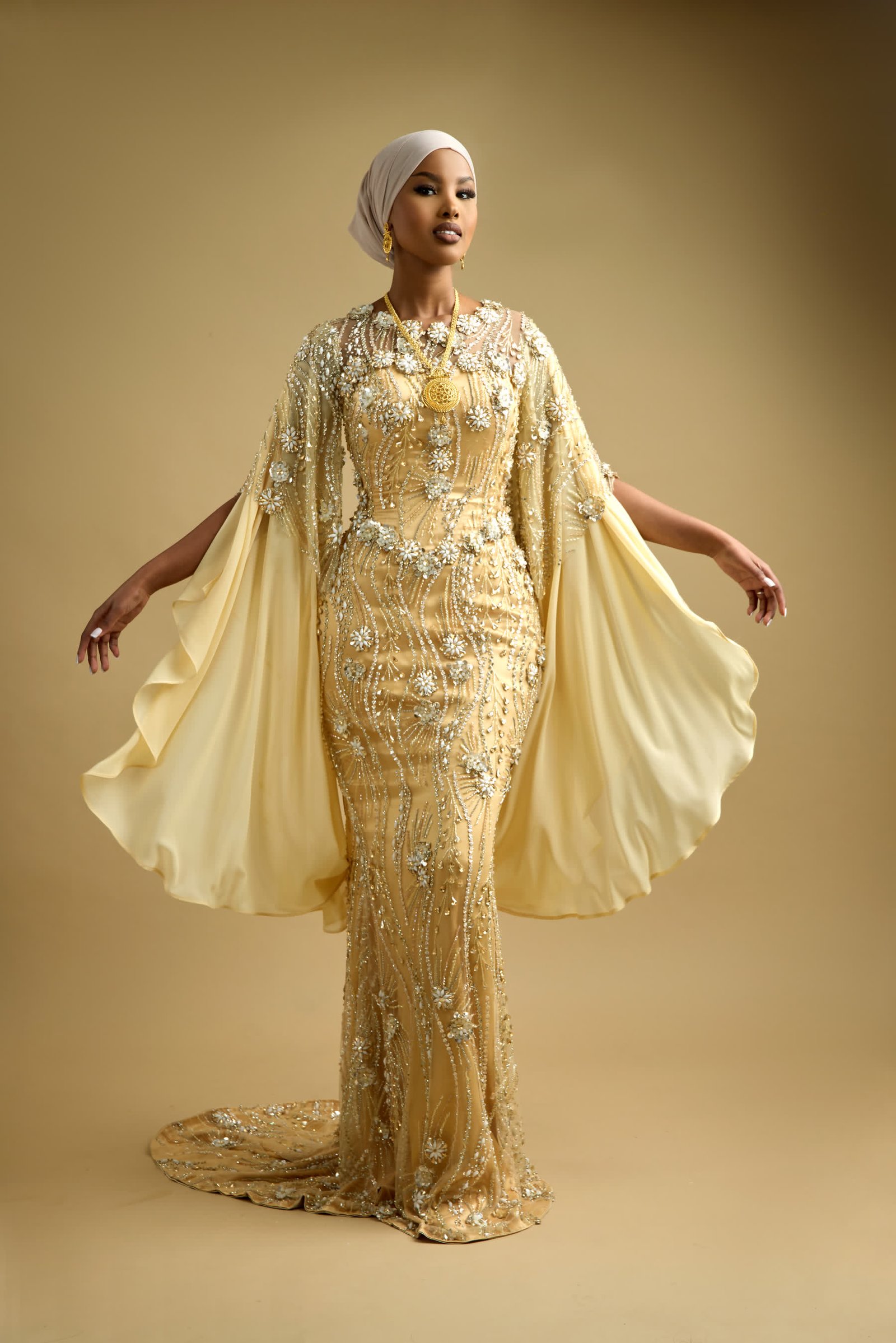 Hand-beaded gold gown photographed mid-movement, the chiffon cape carrying the line of the shoulder